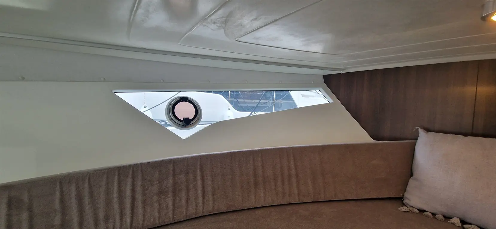 Interior view of a boat, featuring a triangular window and round porthole, with a plush, curved seating area.